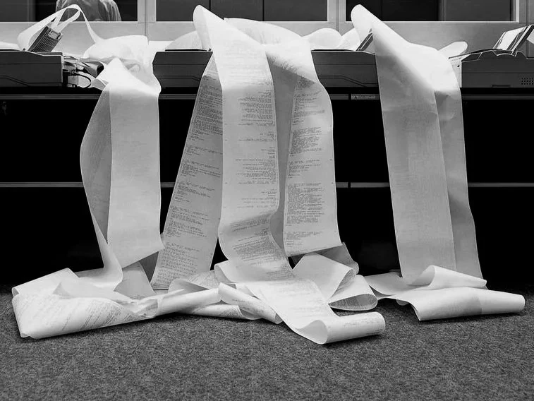 A cluttered office desk with numerous long receipts and papers hanging over the edge onto the floor.