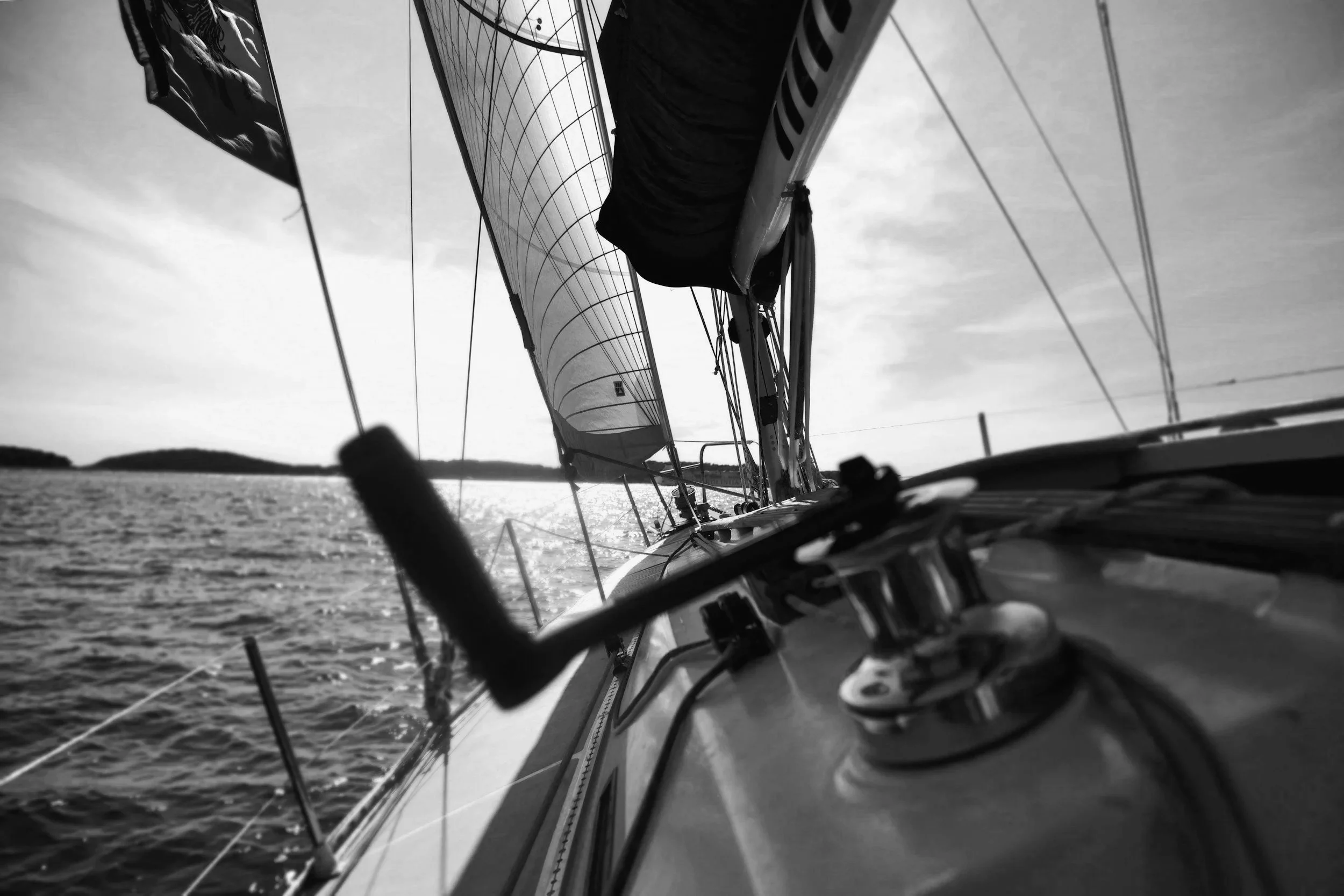 A black and white photo taken from a sailboat looking towards the bow, with the water and distant shoreline in the background.