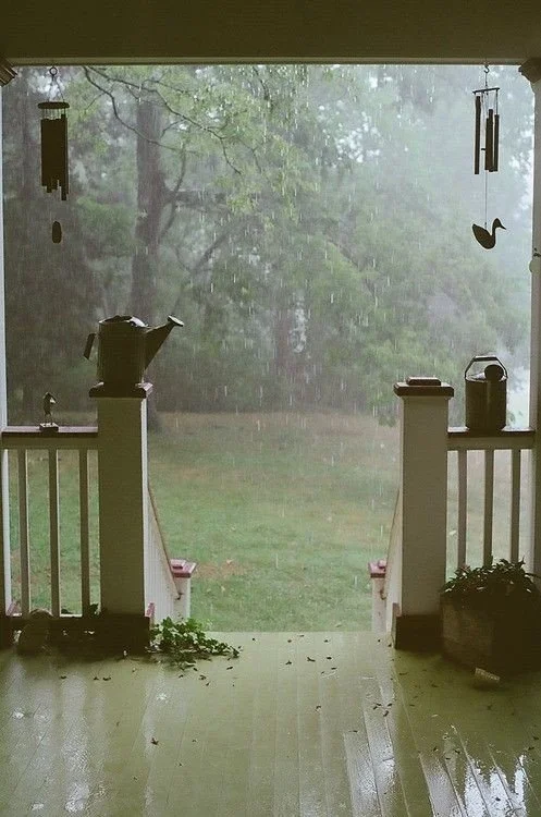 Rainy view from a porch with wind chimes, a teapot, and a kettle, overlooking a grassy yard and trees.