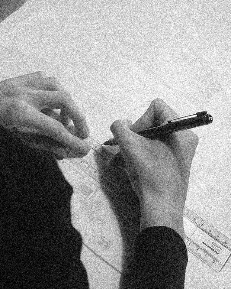 Hands writing on a piece of paper with a pen, on a light-colored desk, with a ruler nearby.