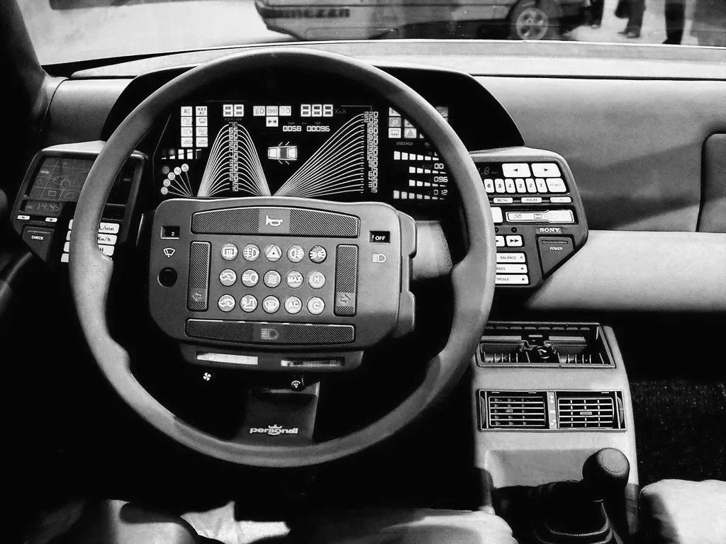 Interior of a vehicle, showing a steering wheel with various control buttons, a digital dashboard display with graphs, and additional control panels with buttons and air vents.
