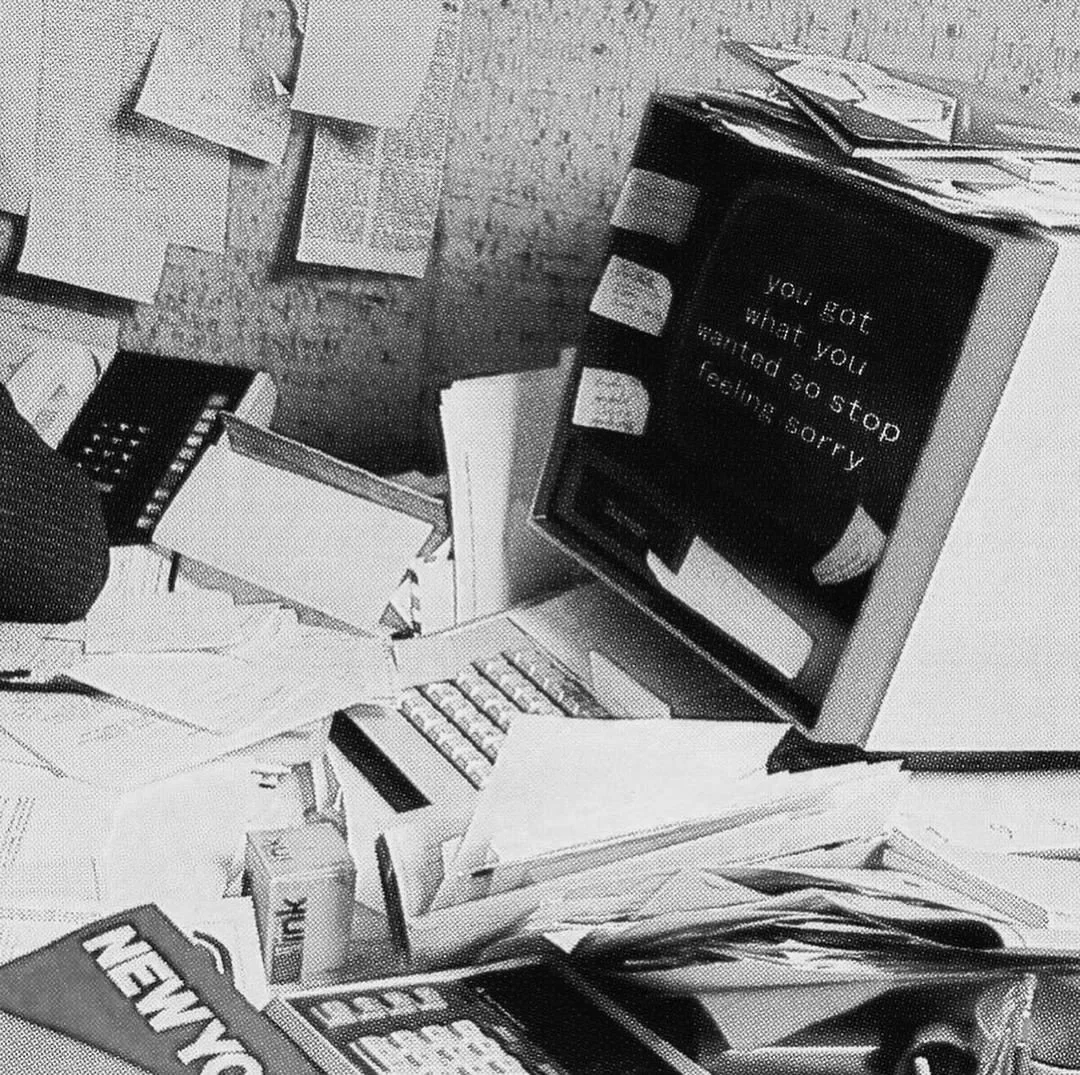 A cluttered desk with papers, a phone, a keyboard, a computer monitor displaying a message, and various office supplies.