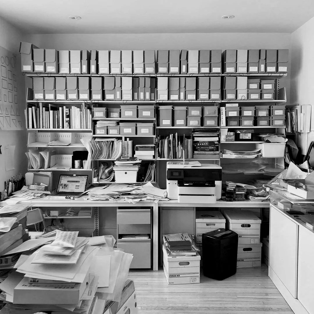 A cluttered office desk with scattered papers, a computer monitor, and a printer in front of a wall filled with shelves holding books, binders, and storage boxes.