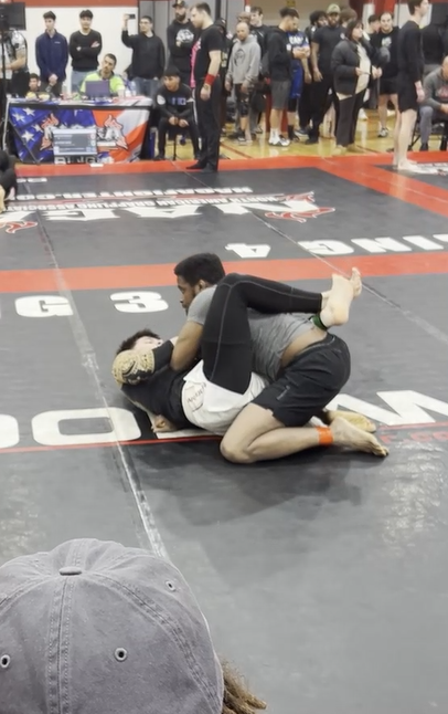 Two men engaged in a Brazilian Jiu-Jitsu match on a black and red mat, with one man on top gripping the other in a grappling hold, while a crowd watches in the background at a competition.