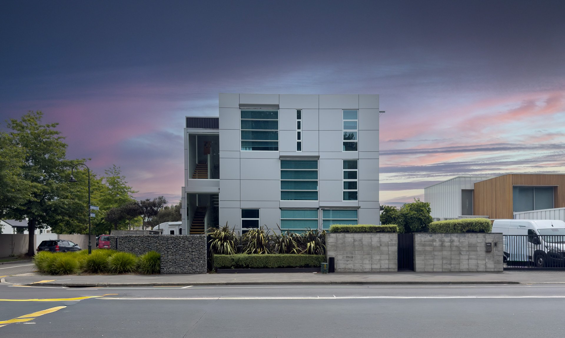 Modern multi-story building with rectangular panels and large windows, surrounded by a Gabion Basket wall and greenery, under a colorful sunset sky.