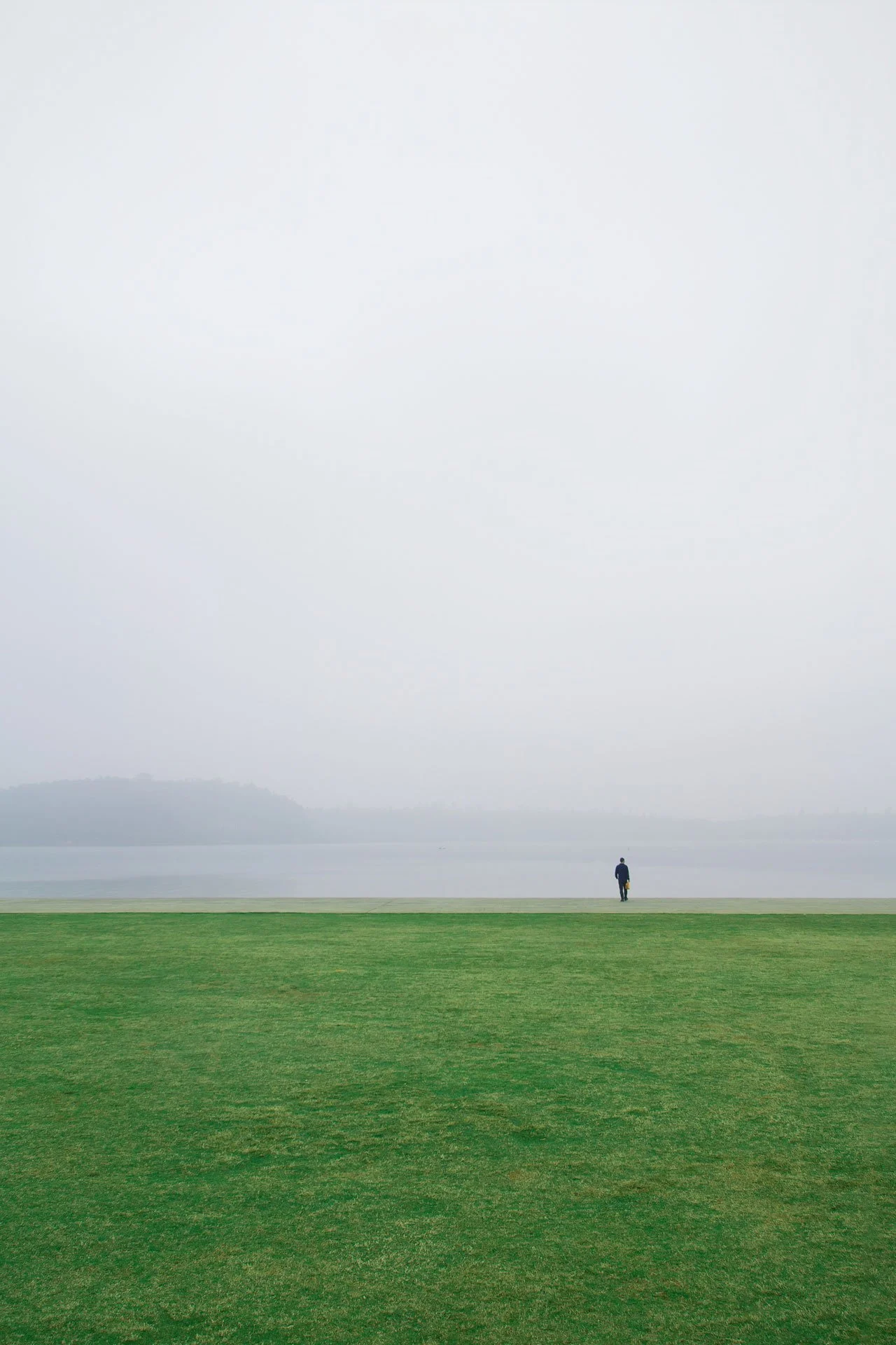 A solitary man walking near a large body of water, with a grassy foreground and overcast sky.