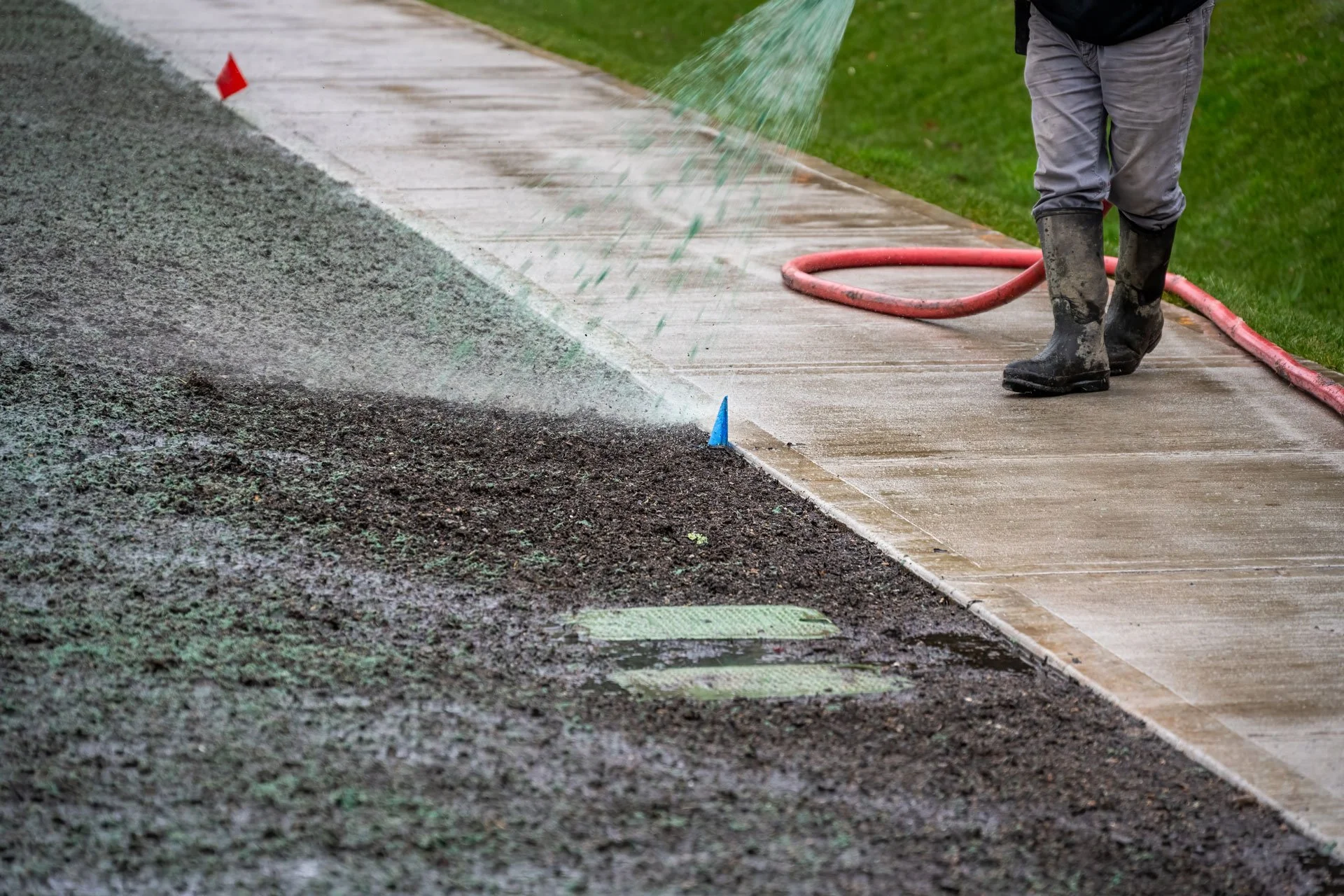A person wearing gray pants and boots waters a wet concrete sidewalk with a hose near a grassy area.