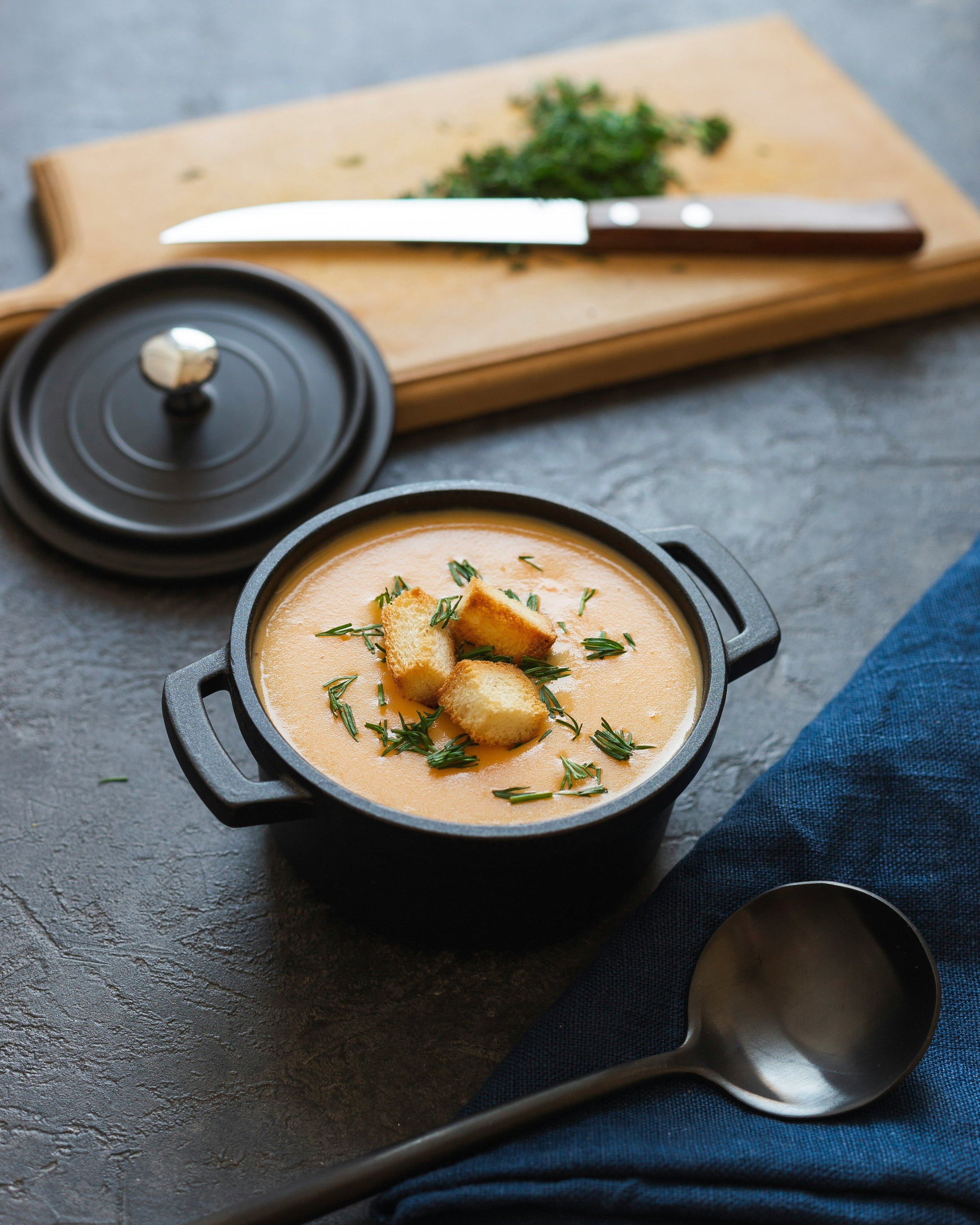A black pot of creamy soup garnished with croutons and herbs, placed on a dark surface next to a blue cloth and silver ladle. In the background, a wooden cutting board with chopped herbs and a knife.