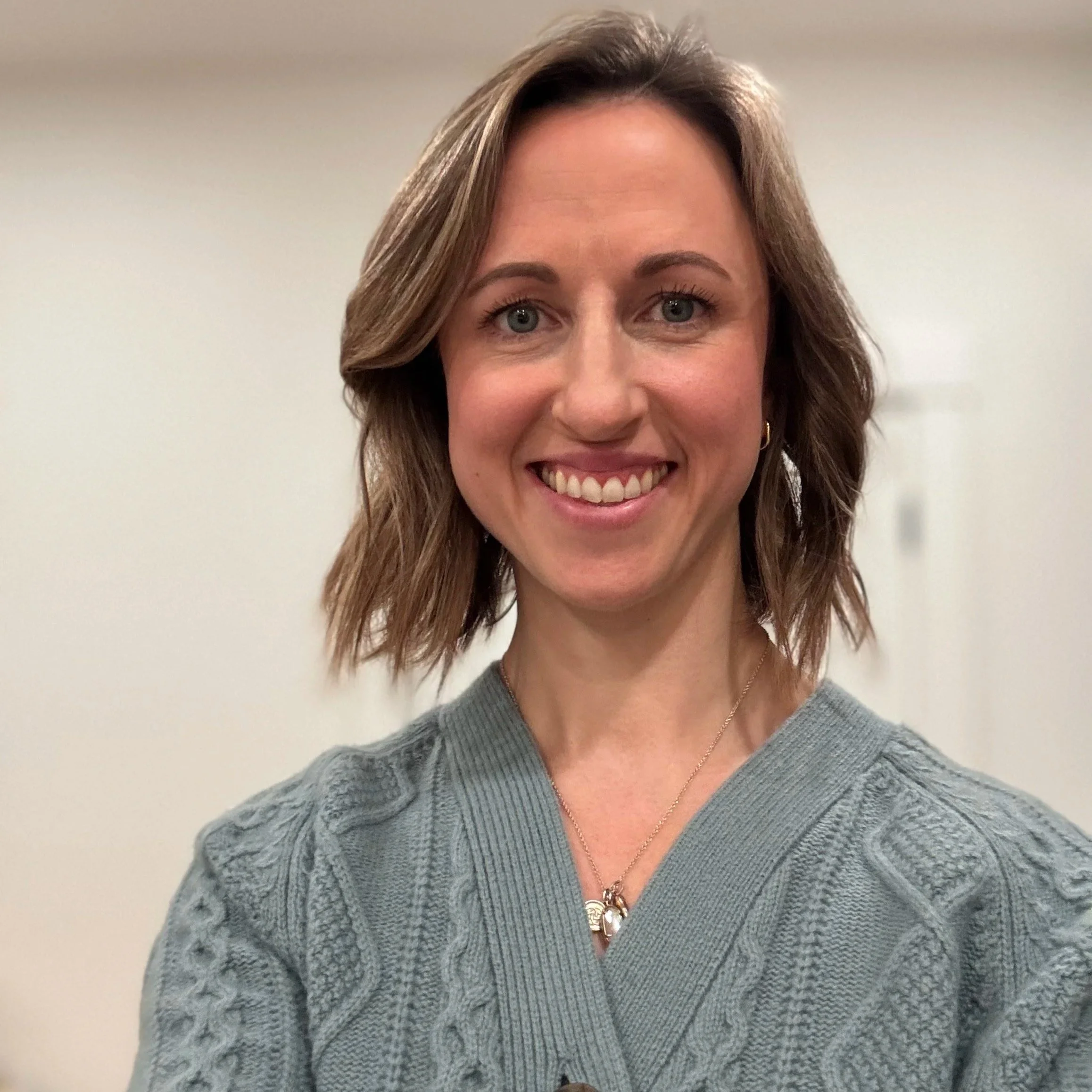 A woman with shoulder-length brown hair, wearing a gray cable-knit sweater and gold jewelry, smiling at the camera against a plain background.