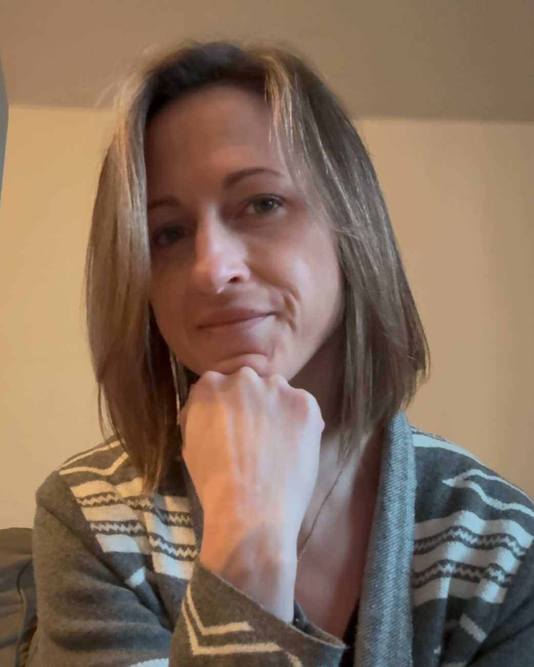 A woman with shoulder-length brown hair, resting her chin on her hand, wearing a patterned sweater, in a room with beige walls.
