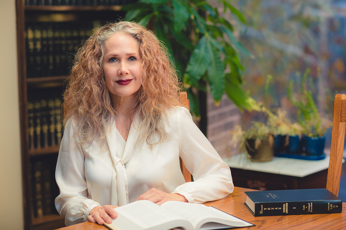 An image of attorney Holly Taylor in a white blouse, seated at a desk with an open law book, with a closed law book on the desk and books and plants in the background.