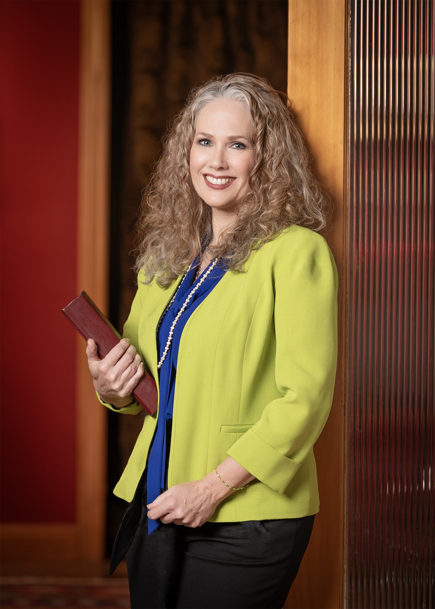 An image of attorney Holly Taylor in a green jacket, leaning against a door frame and holding a red-spined legal book, with a similarly colored wall in the background.