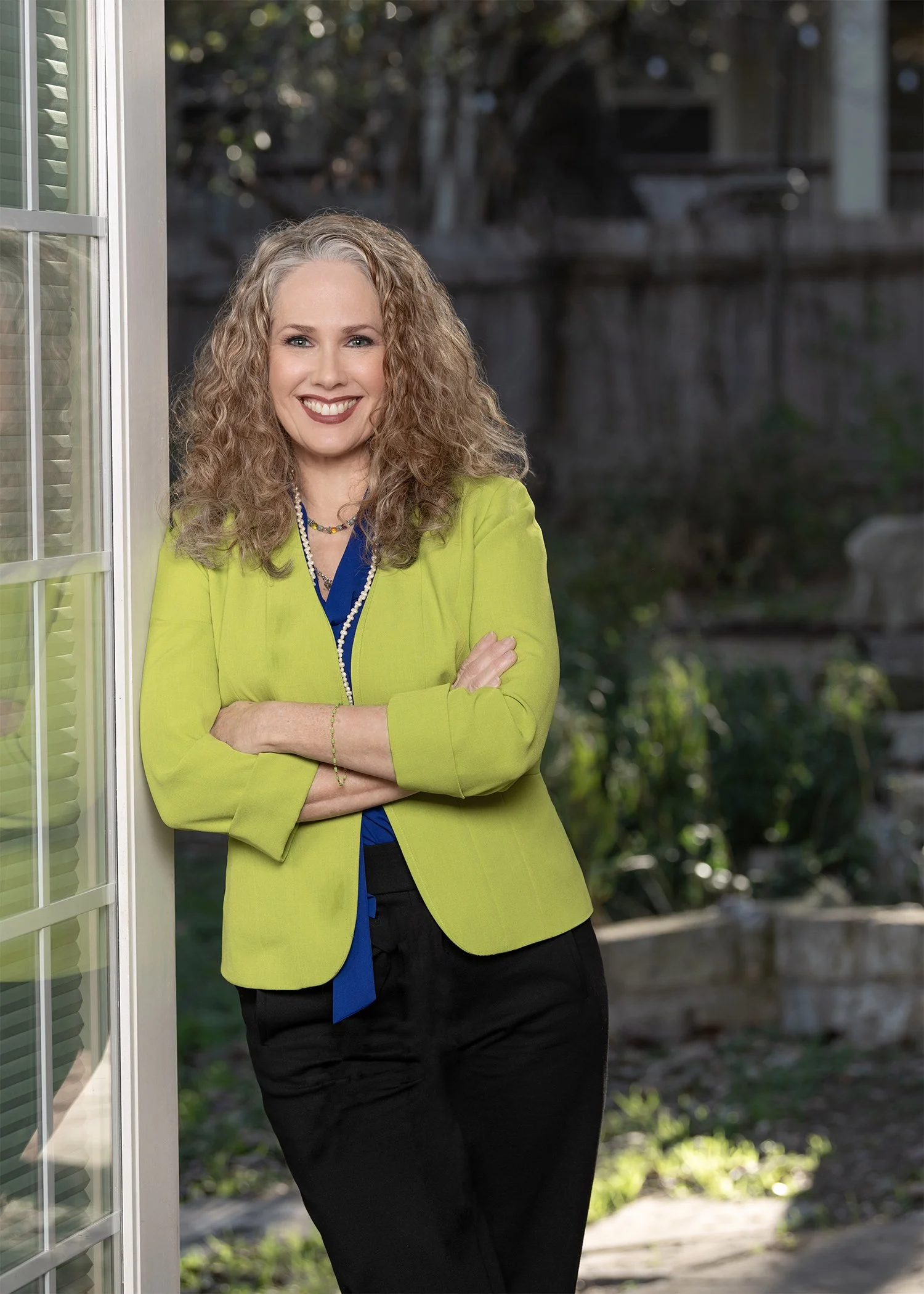 An image of attorney Holly Taylor in a green jacket, leaning against an exterior wall with a garden in the background. Her reflection is visible in a window.