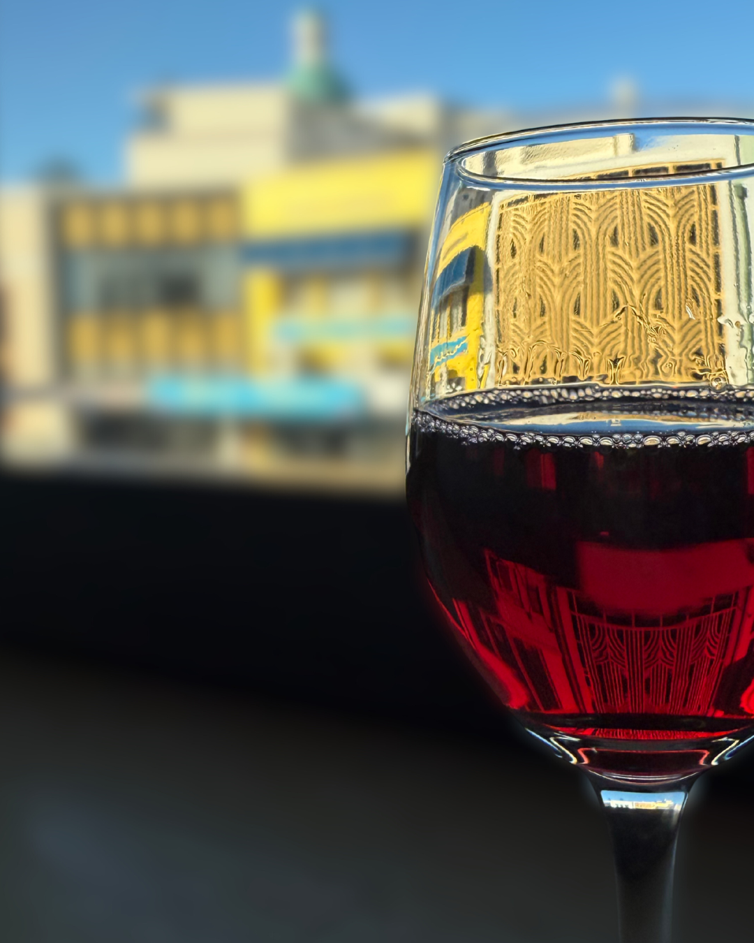 A glass of red wine on a window sill with a blurred cityscape in the background, including buildings and a clear blue sky.
