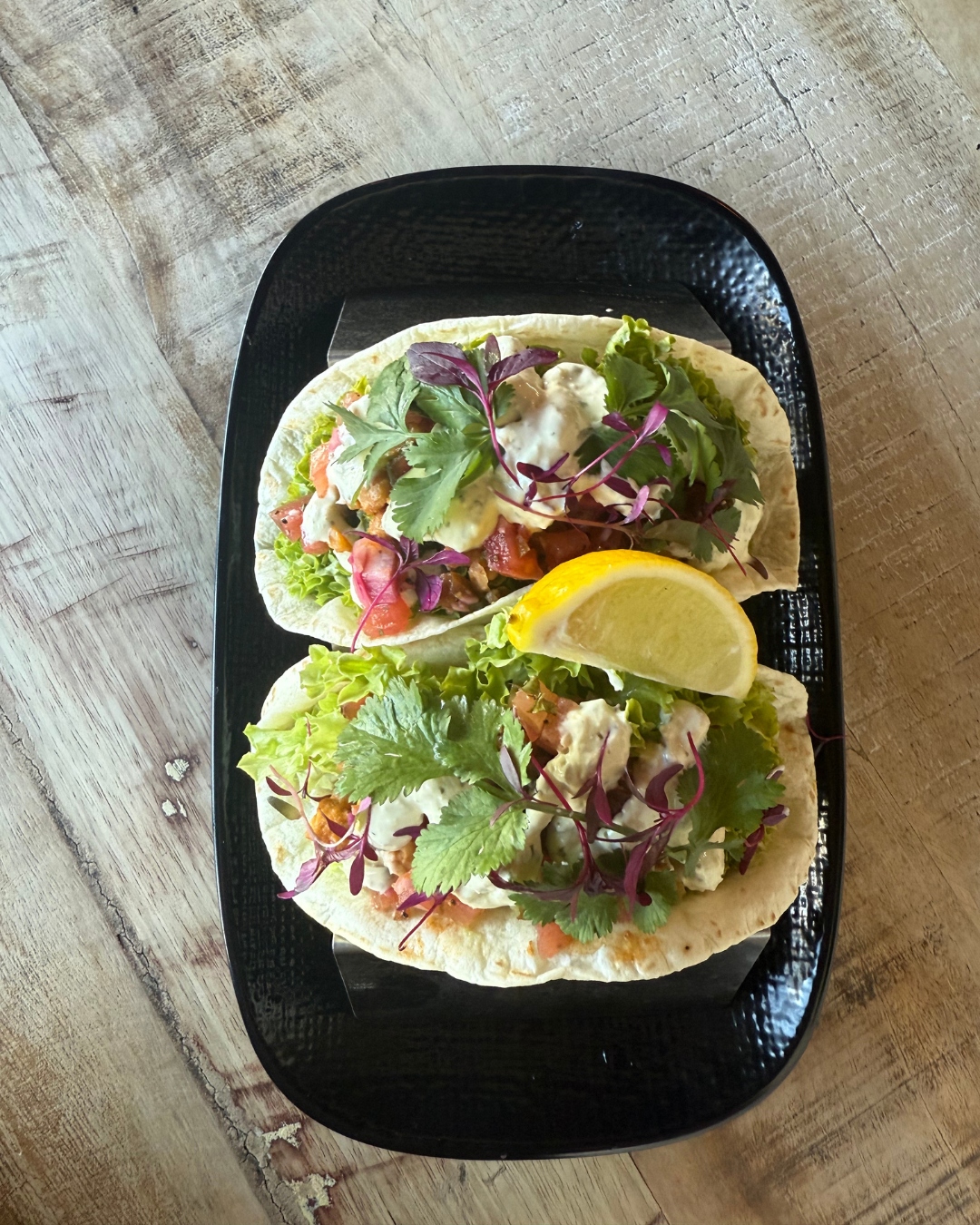Two tacos with lettuce, diced tomatoes, microgreens, and creamy white sauce, garnished with lemon wedges on a black plate.