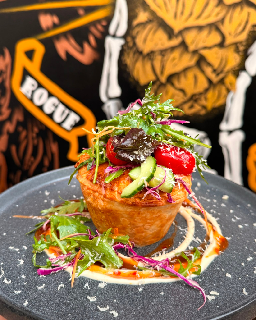 A salad served in a small fried tortilla bowl, topped with mixed greens, cherry tomatoes, cucumber slices, and shredded cheese, on a black plate with sauce and shredded cheese garnish.