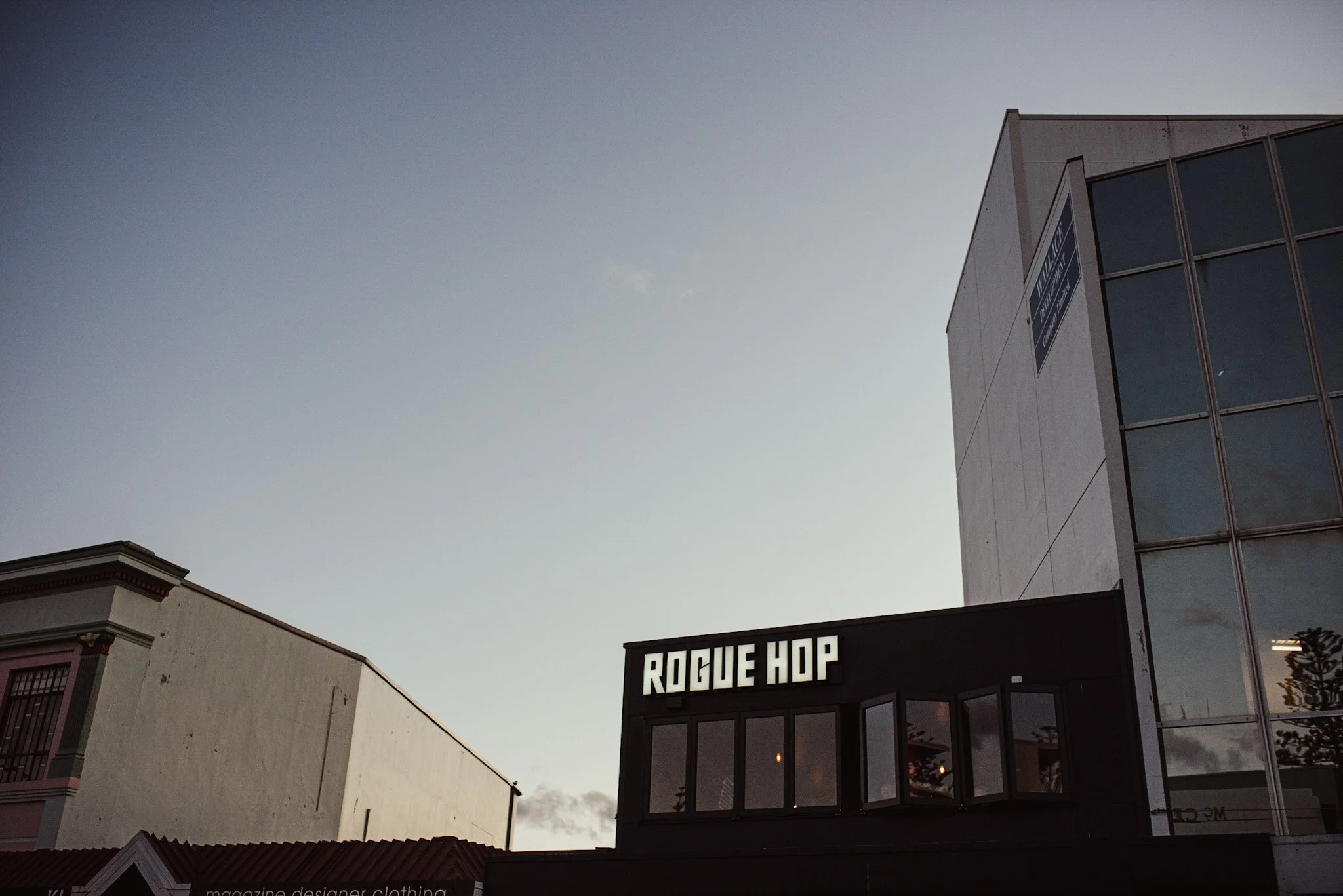 City street scene showing a modern building with large windows, a sign reading 'ROGUE HOP', and a traditional-style building with decorative trim, under a clear sky.