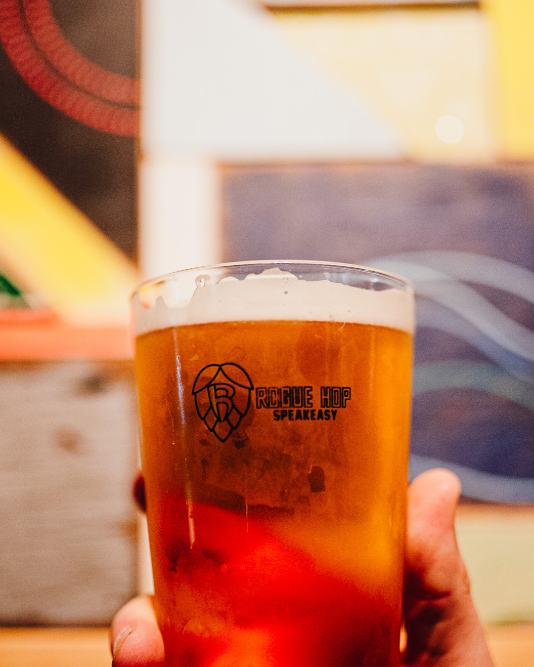 A glass of beer with a logo that says Rogue Hop Speak Easy, held in a person's hand in an indoor setting.