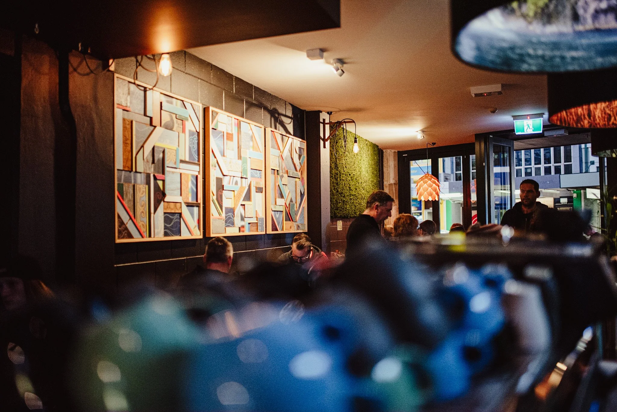 The image depicts the interior of a lively restaurant or cafe with dim lighting and a modern decor style. There are several people seated at tables, engaged in conversation or dining. On the wall, there is a colorful abstract artwork made of various geometric shapes and patterns. The scene is illuminated by warm light fixtures hanging from the ceiling and wall-mounted lights, creating a cozy atmosphere. Large windows at the back reveal an urban street scene outside.