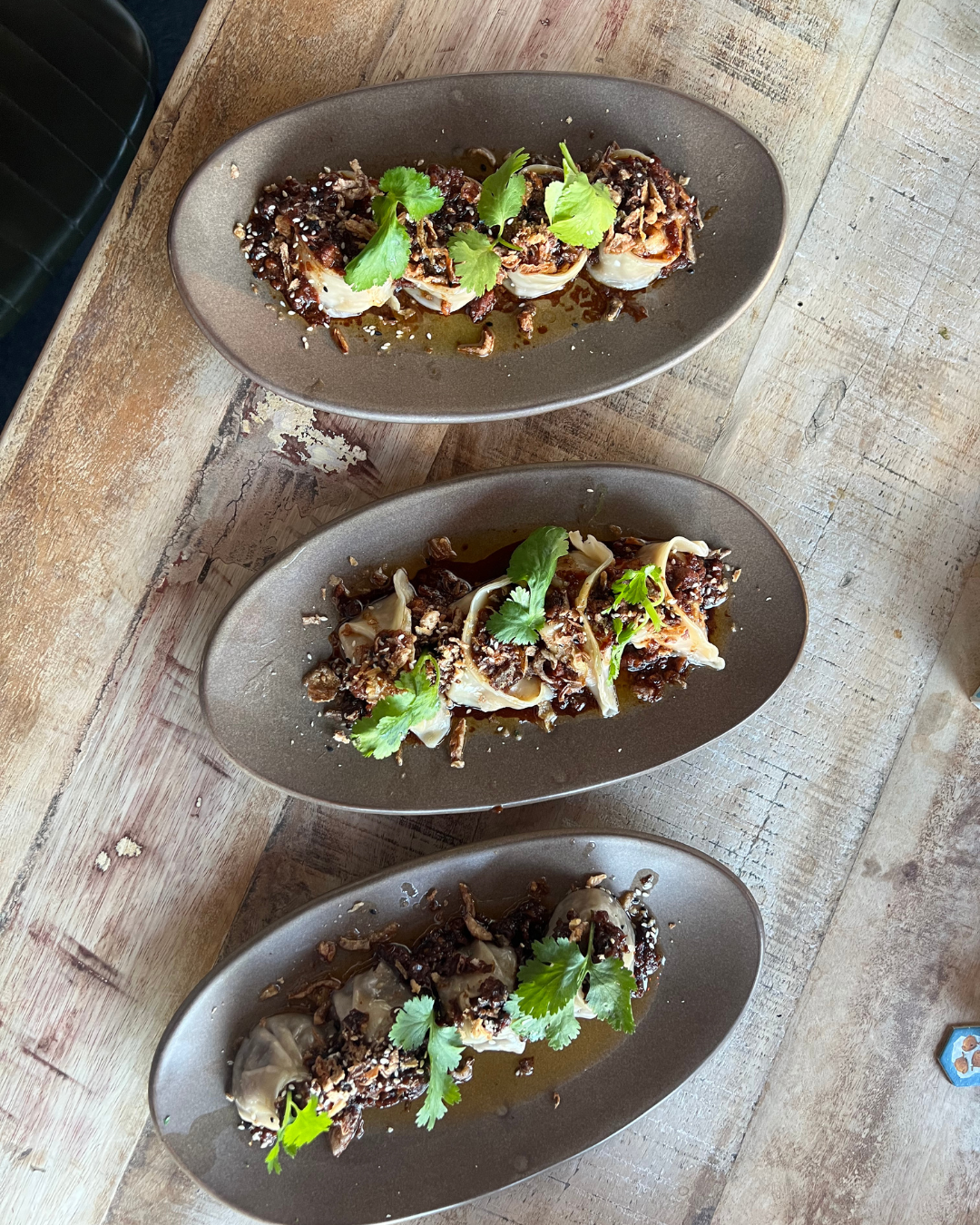 Three oval plates with Asian dumplings topped with cilantro, sesame seeds, and a dark sauce, placed on a rustic wooden table.