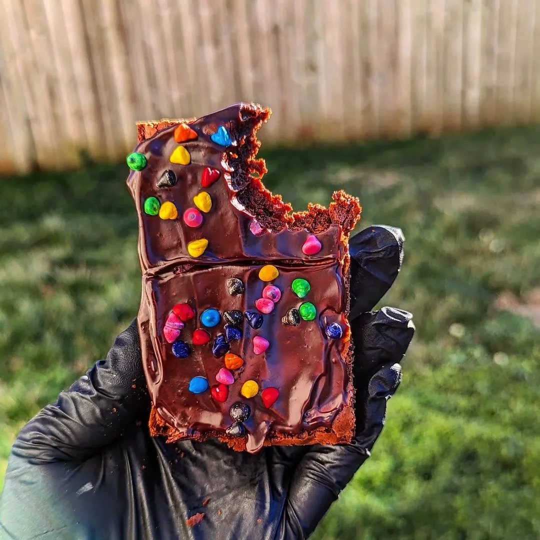 Hand in black glove holding a partially eaten chocolate brownie with colorful sprinkles outdoors, with a blurred wooden fence and grass in the background.