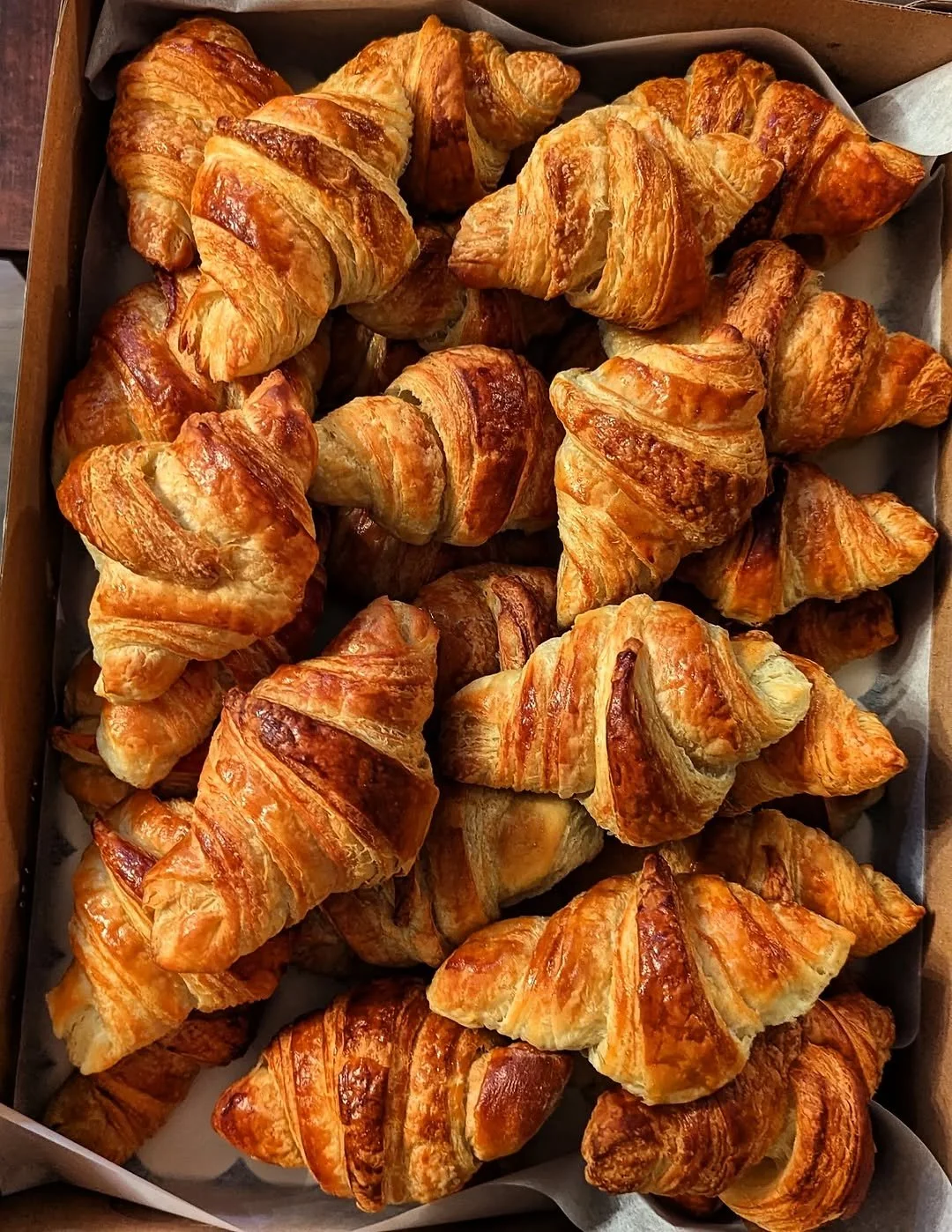 A tray filled with freshly baked, golden-brown croissants, arranged in a cardboard box lined with parchment paper.