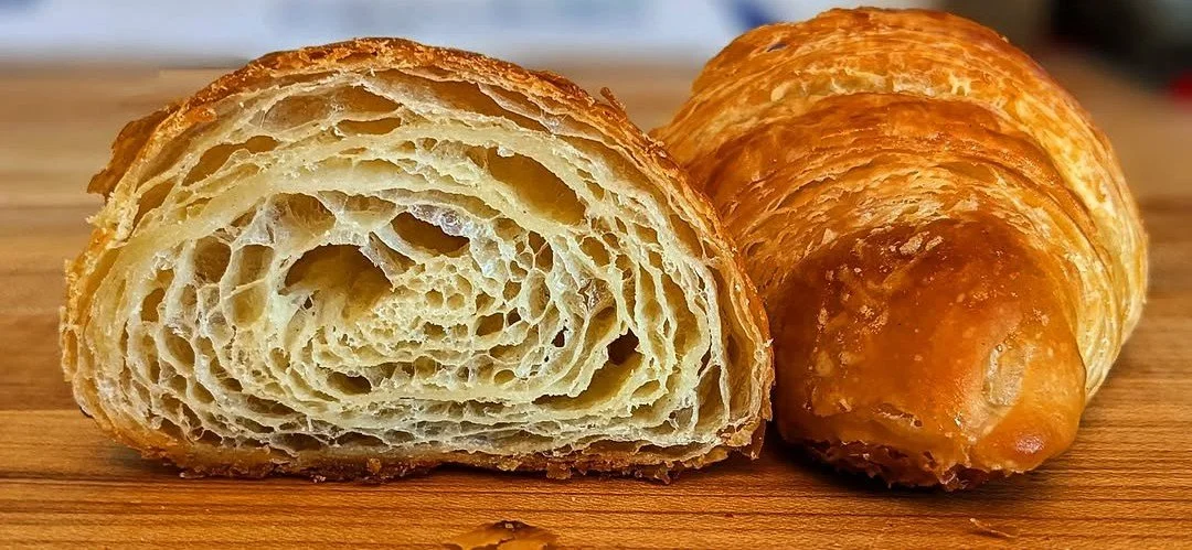 Close-up of a flaky croissant with a crispy golden-brown exterior, cut in half to show its layered, airy interior, placed on a wooden surface.
