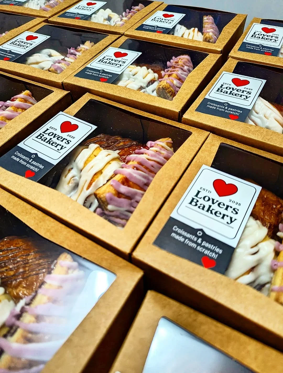 Multiple boxes of baked goods from Lover's Bakery, each with a clear top displaying assorted croissants and pastries with icing, labeled with Lover's Bakery branding and description.