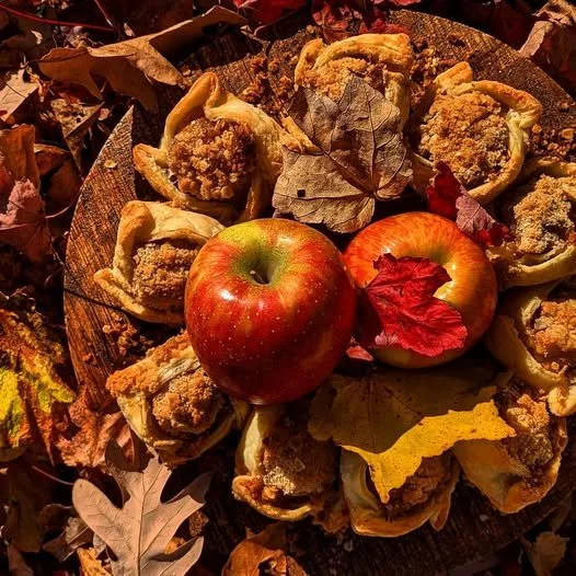 Two red apples surrounded by baked stuffed bell peppers with crumble topping, placed on a wooden surface outdoors amid fallen autumn leaves.