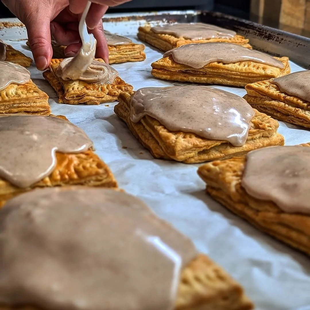 A baker uses a piping bag to drizzle icing on rectangular apple turnovers topped with icing on a baking sheet lined with parchment paper.