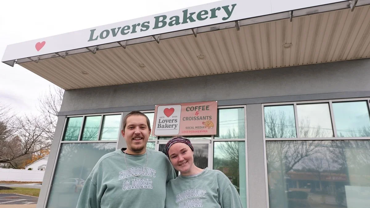 Two people smiling outside Lovers Bakery, wearing matching gray shirts that say 'Croissant Slinger'.