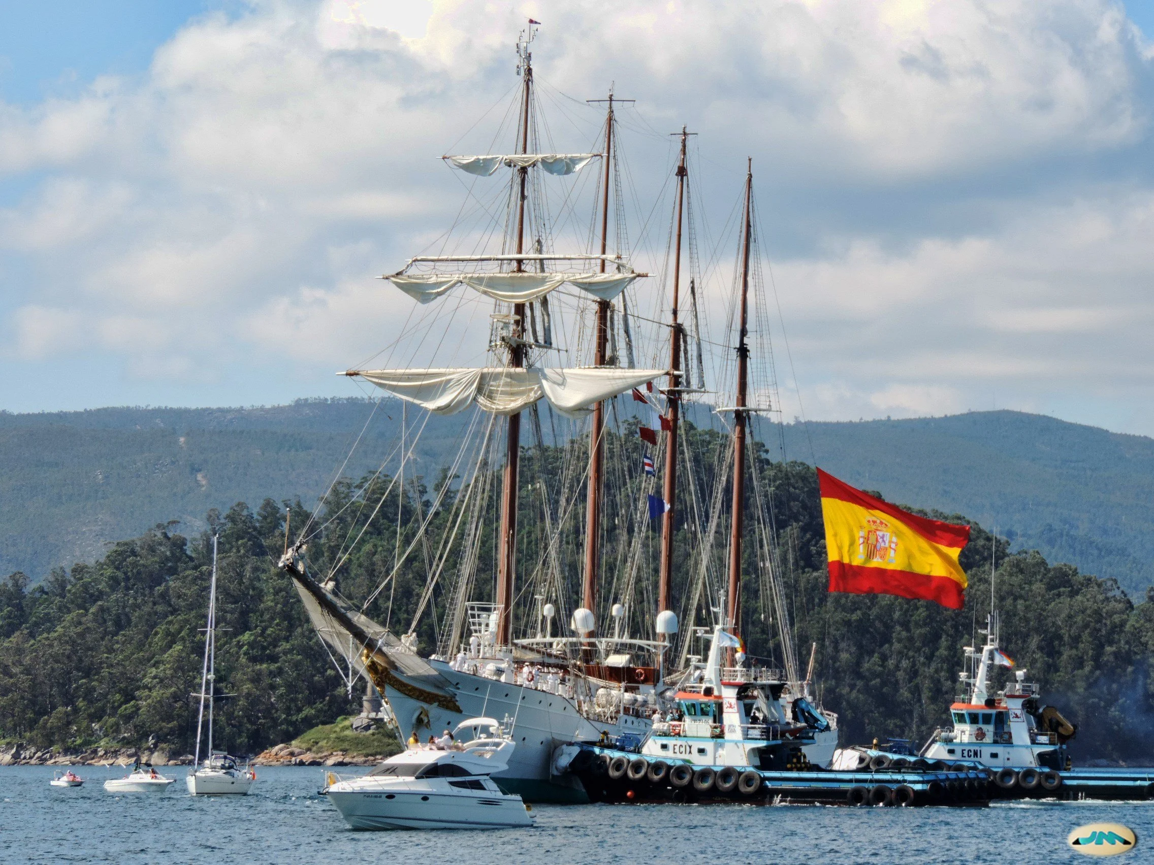 A large sailing ship with three masts, flying a Spanish flag, anchored near smaller boats in a bay with forested hills in the background.