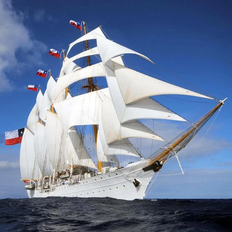 A large sailing ship with white sails and multiple red, white, and blue flags sailing on the ocean under a clear blue sky.