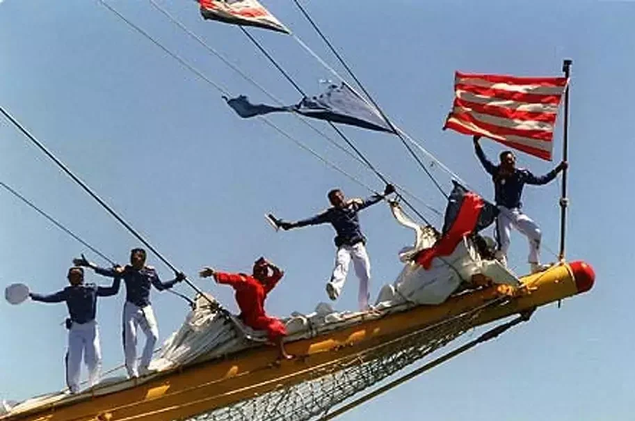 People dressed as sailors holding American flags on a ship's mast, with a clear blue sky in the background.