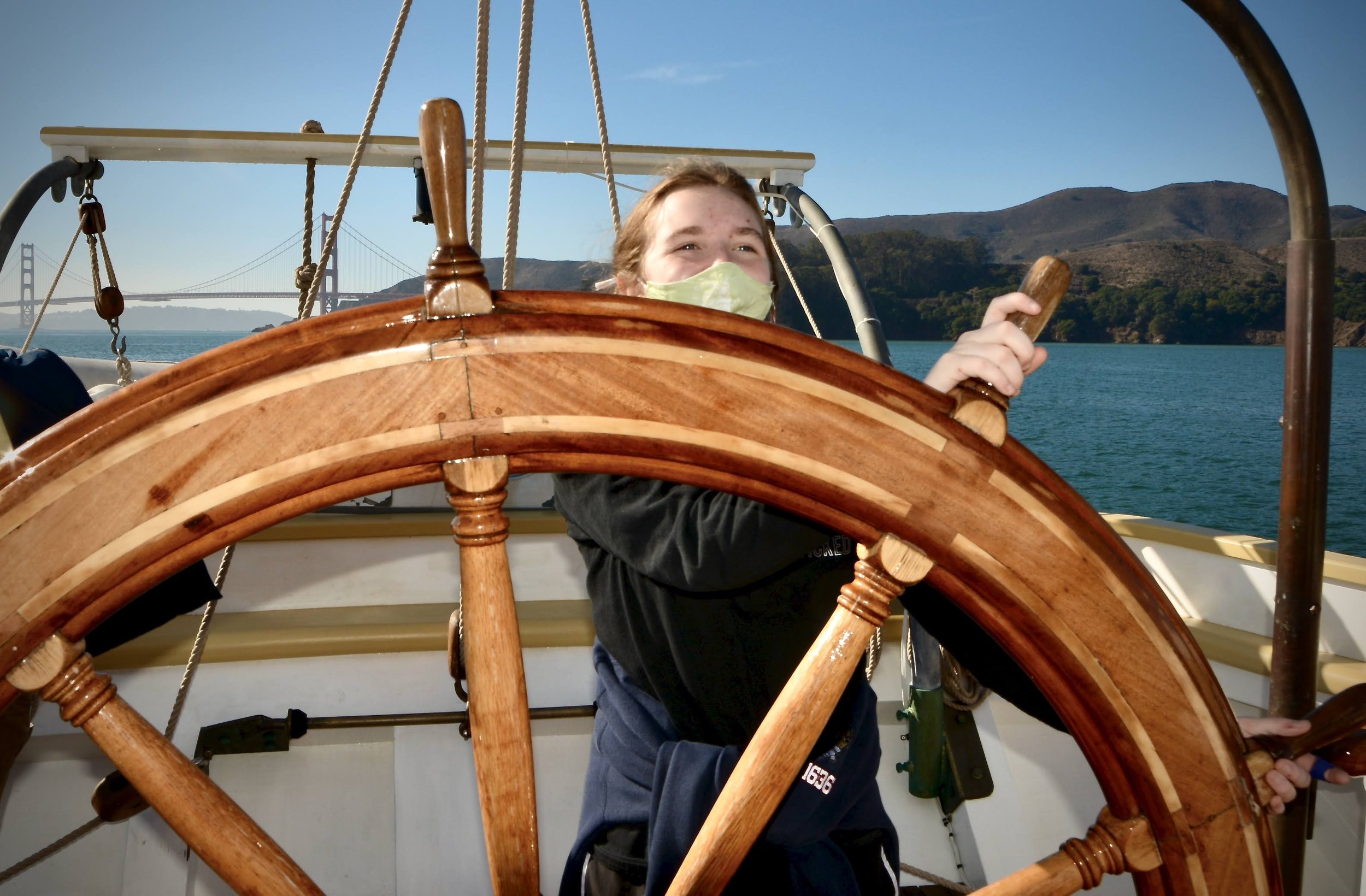 A young woman wearing a face mask steering a sailboat, with a view of water, hills, and a bridge in the background.