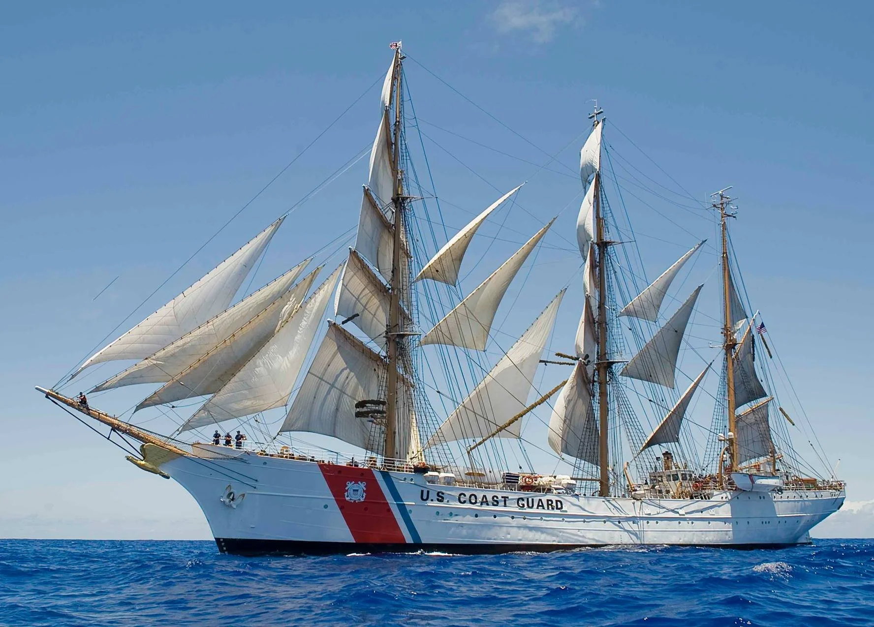A U.S. Coast Guard tall ship with multiple sails on the ocean under a blue sky.
