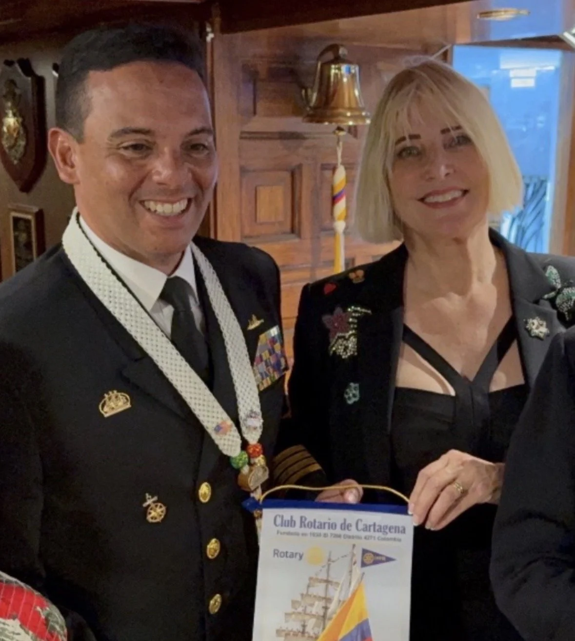 A man in a formal military uniform with medals and a woman in a black top and blazer with embroidered pins, holding a Rotary Club banner, smiling indoors with wooden walls and a bell in the background.