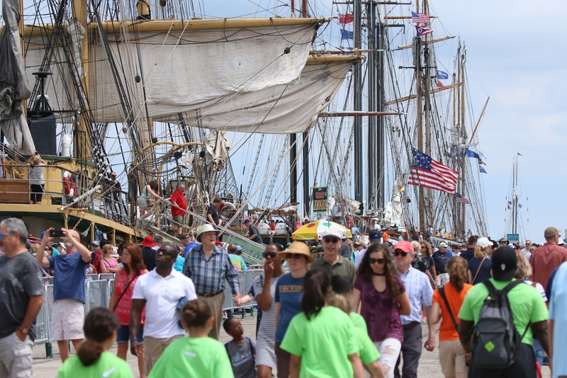 A crowded pier with multiple large sailing ships docked, tall masts, and American flags, filled with people including families, tourists, and crew members, some taking photos and enjoying the scene.