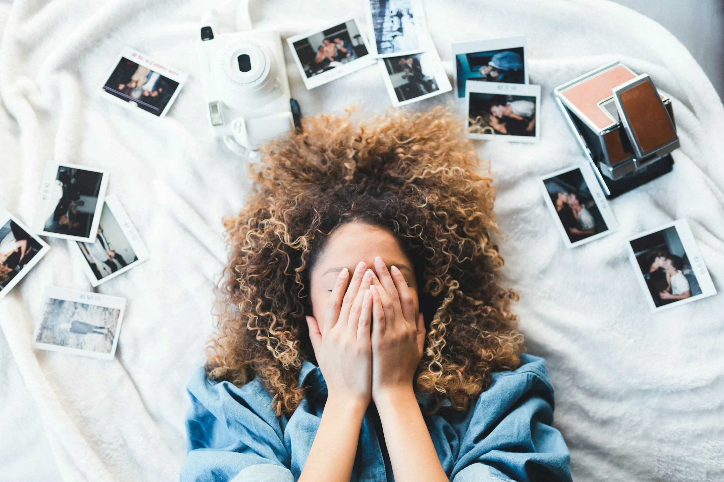 A woman with curly hair lying on a white blanket, covering her face with her hands, surrounded by instant photo prints, a stack of makeup palettes, a camera, and a few personal items.