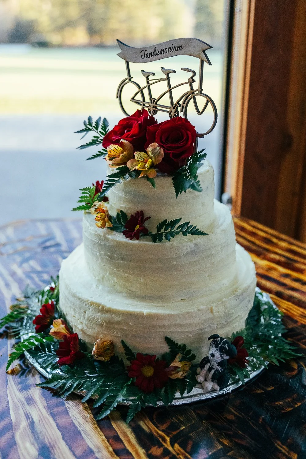 A three-tiered white wedding cake decorated with red roses, yellow flowers, green leaves, a bicycle top cake topper with a banner reading 'Tandemonium', and a small dog figurine at the base.