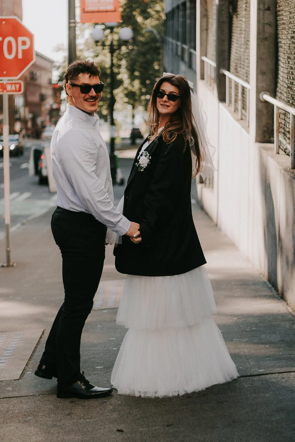 A couple holding hands and smiling outdoors on a city sidewalk, with a man wearing sunglasses, a white shirt, and black pants, and a woman in a wedding dress with a black jacket, both wearing sunglasses.