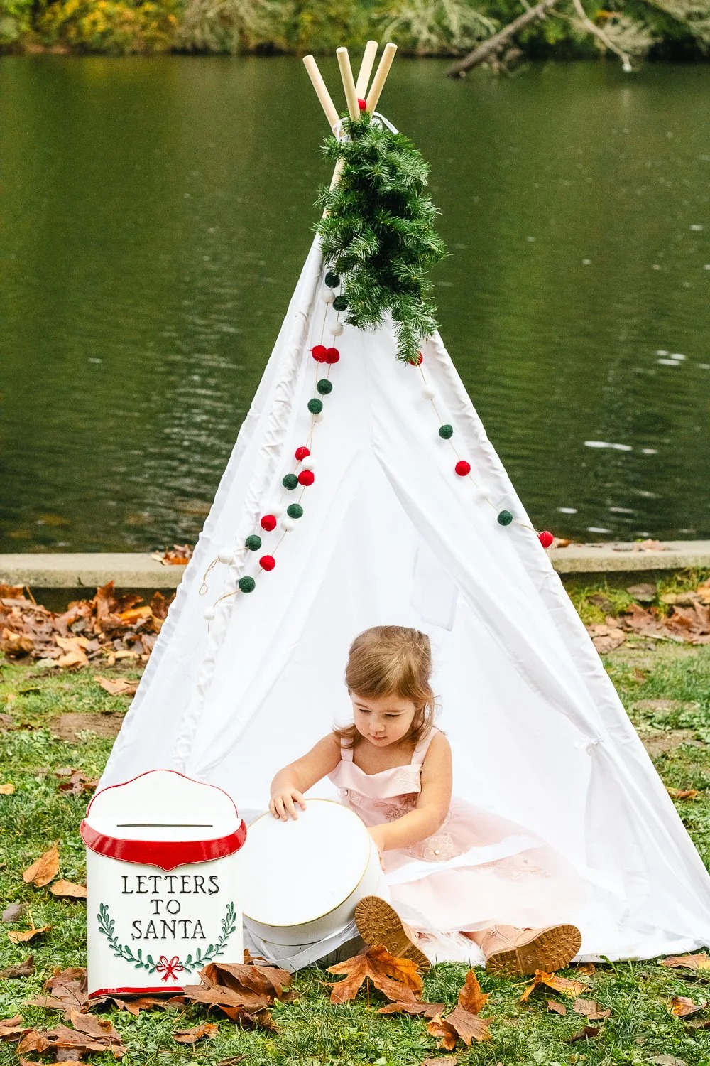 A young girl in a pink dress sitting inside a white teepee decorated with green and red Christmas ornaments, near a lake with autumn leaves on the ground, and a white box labeled "Letters to Santa" in front of her.
