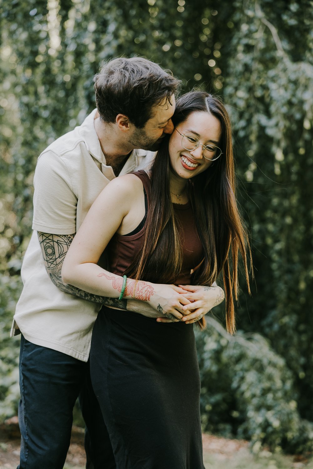 A couple sharing a happy moment outdoors, with the man kissing the woman's cheek and the woman smiling, surrounded by greenery.