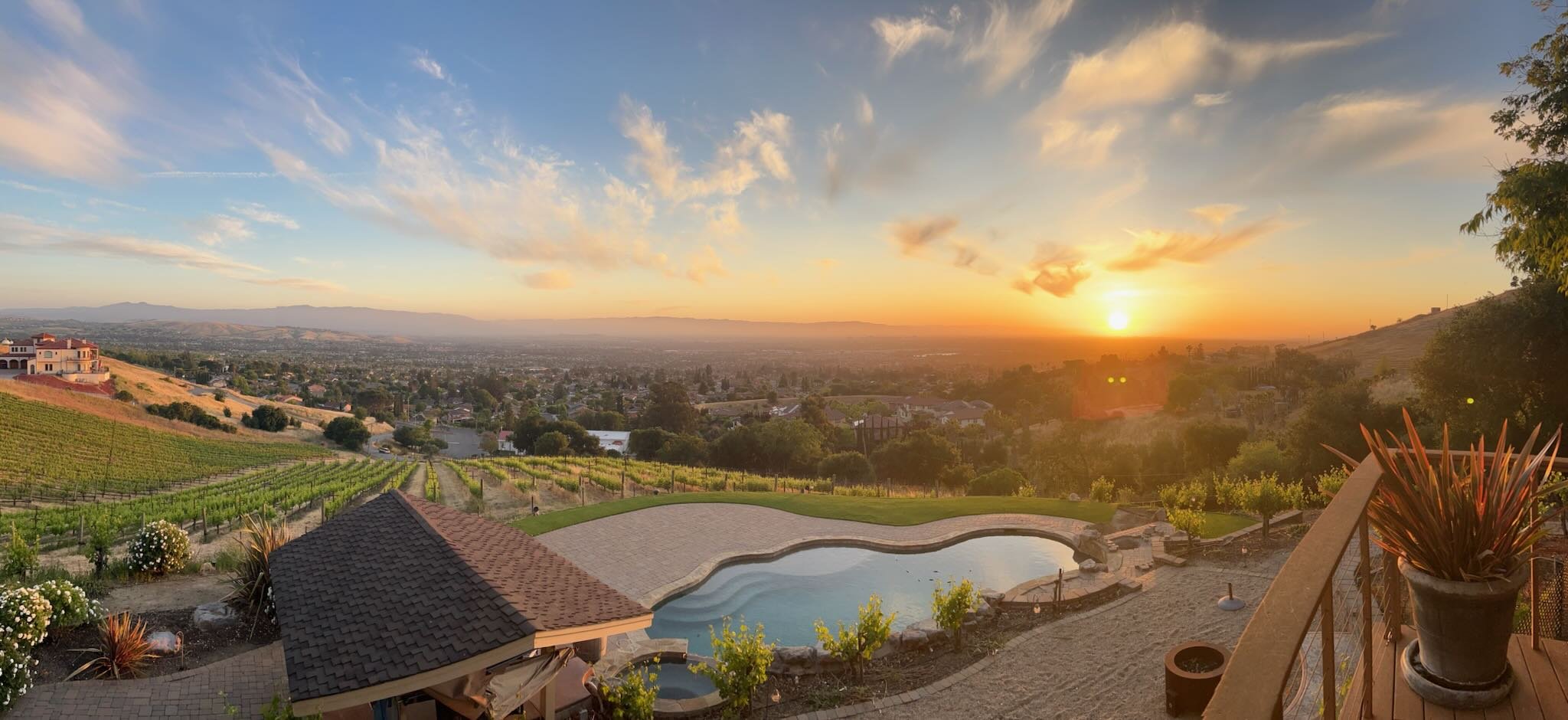 A scenic view of a sunset over a hilly landscape with vineyards, residential houses, and a swimming pool in the foreground.