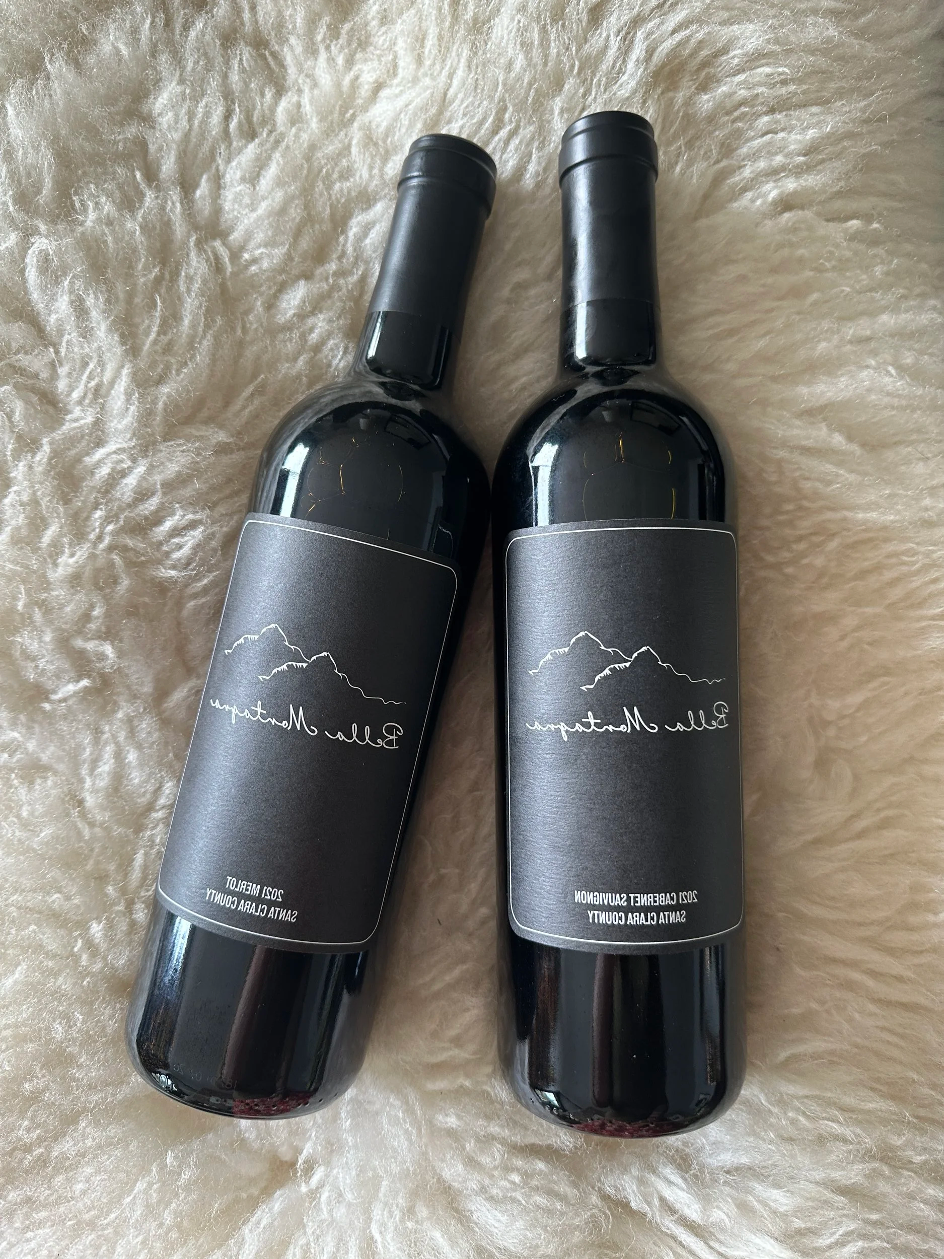 Two bottles of red wine with black labels and caps, placed on a beige fuzzy surface.