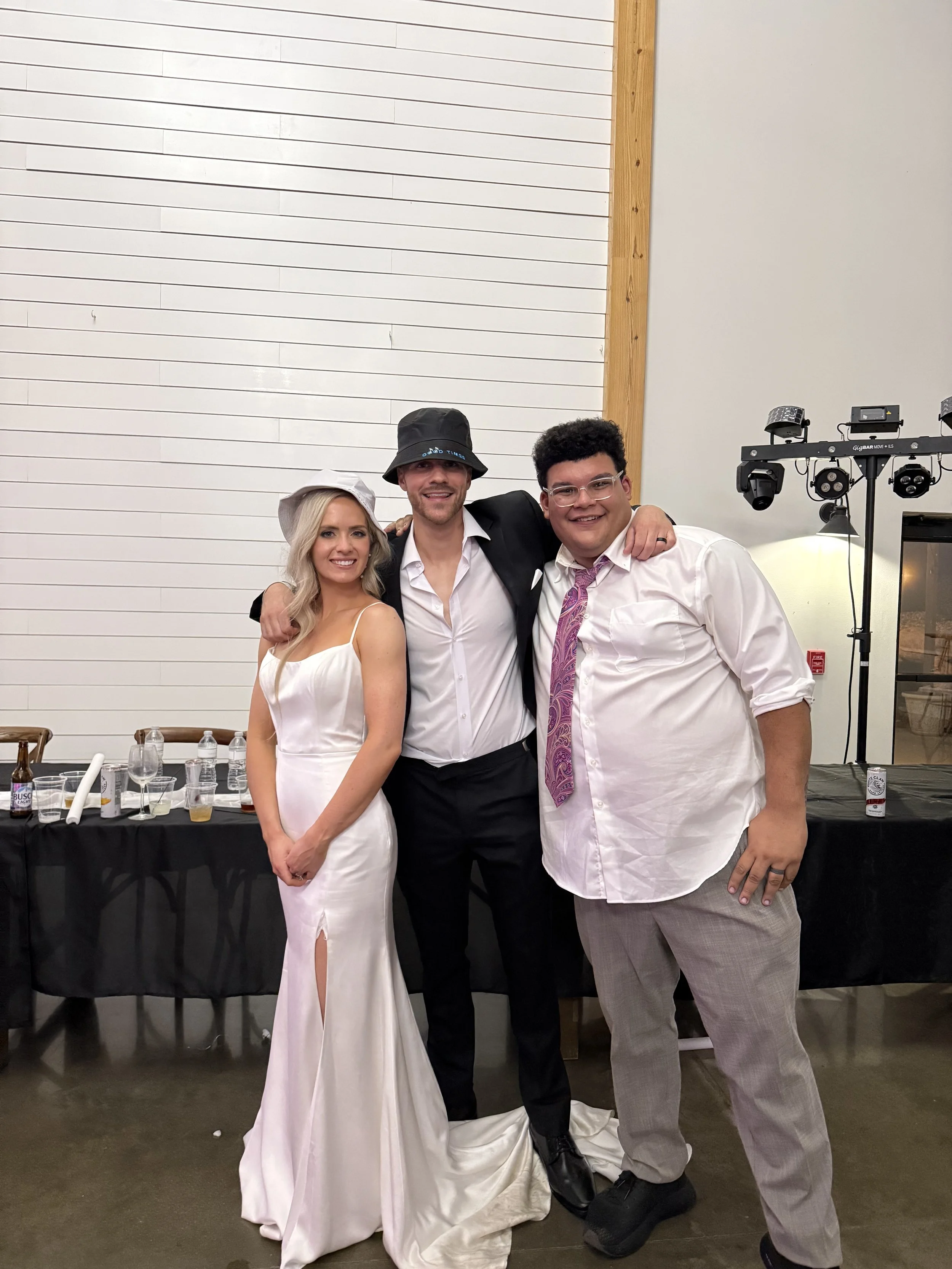 Three people smiling and posing at a party or celebration, with drinks and equipment in the background.