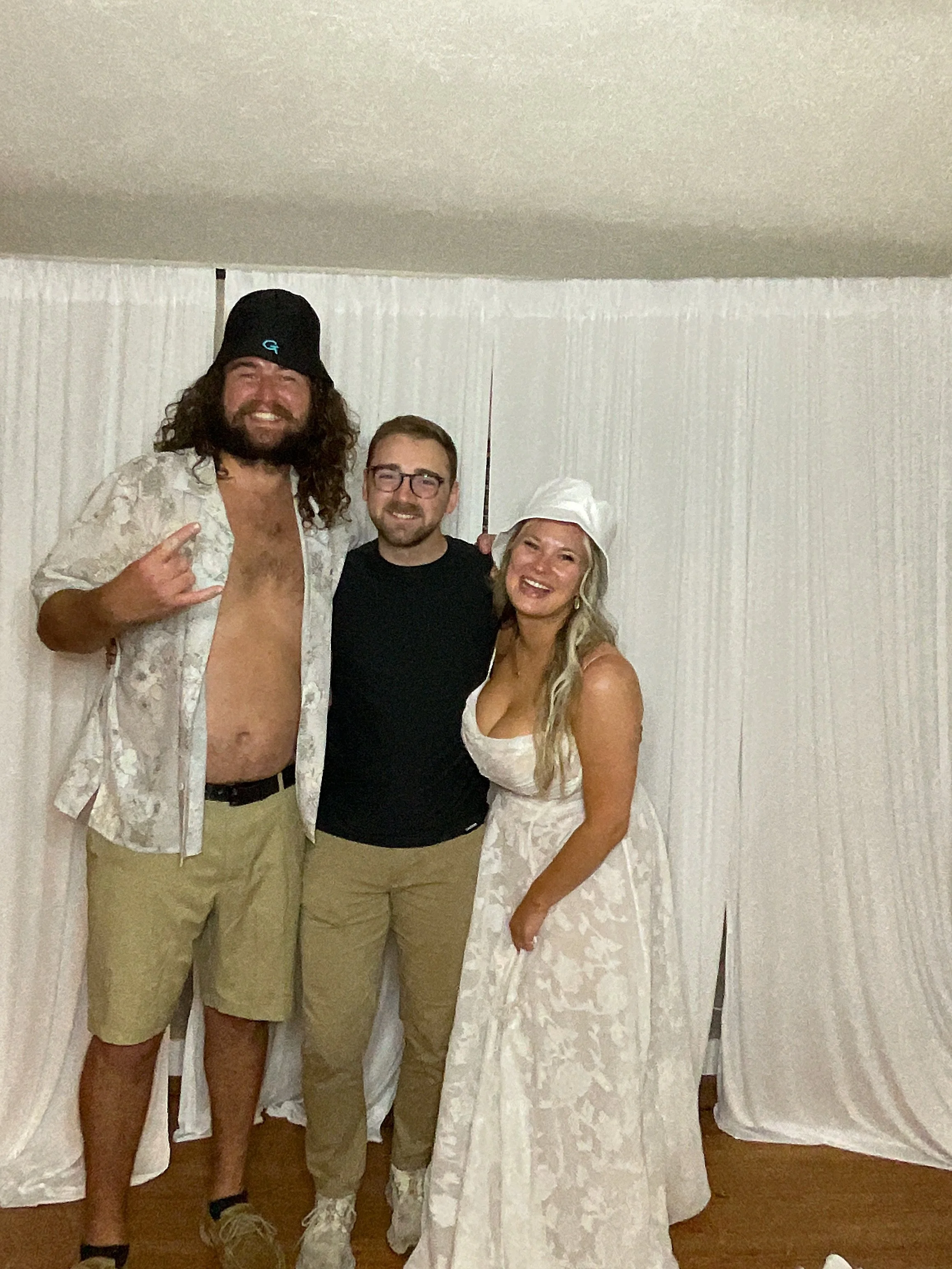 Three people smiling and posing for a photo indoors. The person on the left is tall with long curly hair, wearing a black beanie, open floral shirt, and khaki shorts. The person in the middle has short hair, glasses, and a black shirt. The person on 