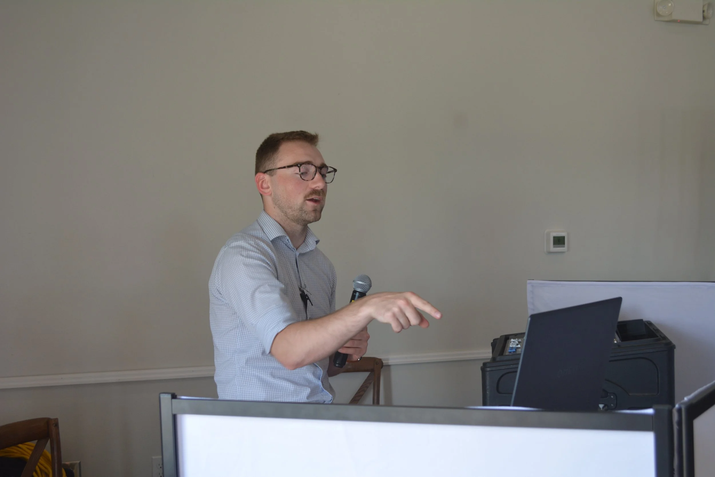 A man wearing glasses and a light-colored checkered shirt standing and holding a microphone, pointing at a laptop on a table in a room with plain walls.
