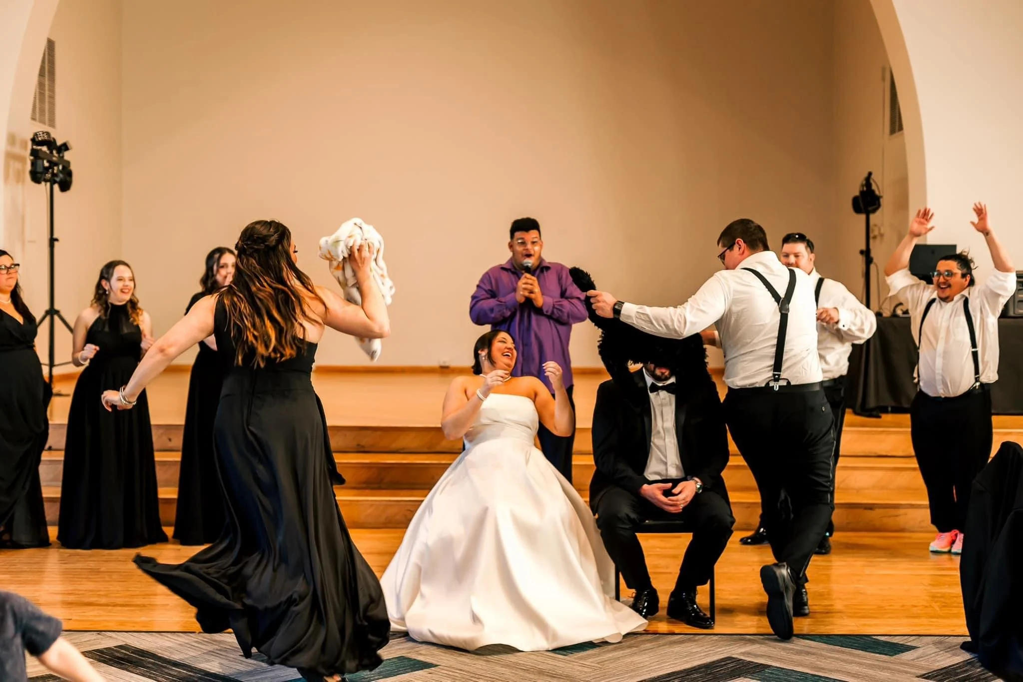 Wedding reception scene with bride in white gown sitting on a chair, groom in tuxedo, wedding party dancing and celebrating on stage, one person in purple jacket singing into microphone, others in black dresses and white shirts with suspenders, joyfu