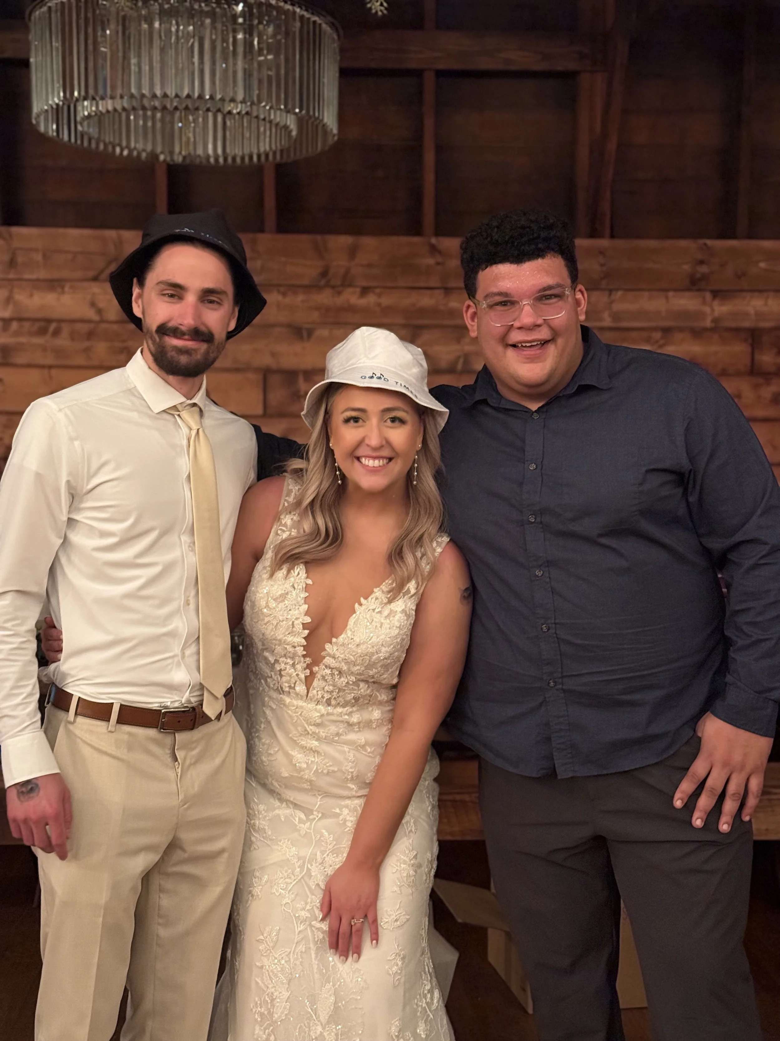 Three people are posing indoors in front of a wooden wall. The person on the left is a man with a beard wearing a white shirt, a beige tie, beige pants, and a black hat. The woman in the middle is wearing a white wedding dress and a white bucket hat 